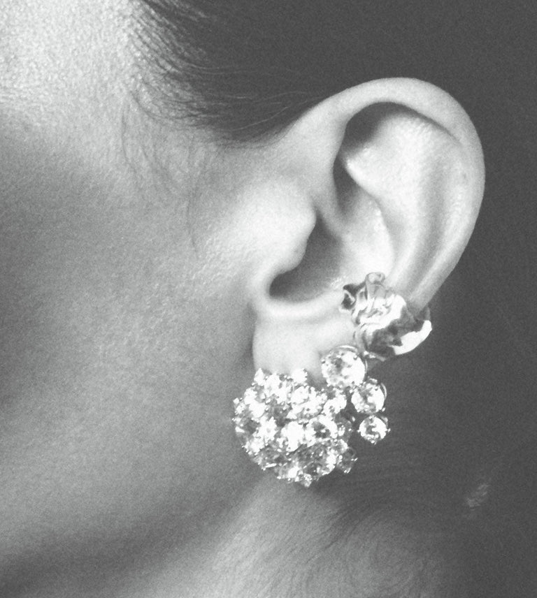 A close-up black and white photograph of a person's ear wearing a large, sparkling cluster stud earring with multiple clear gemstones.